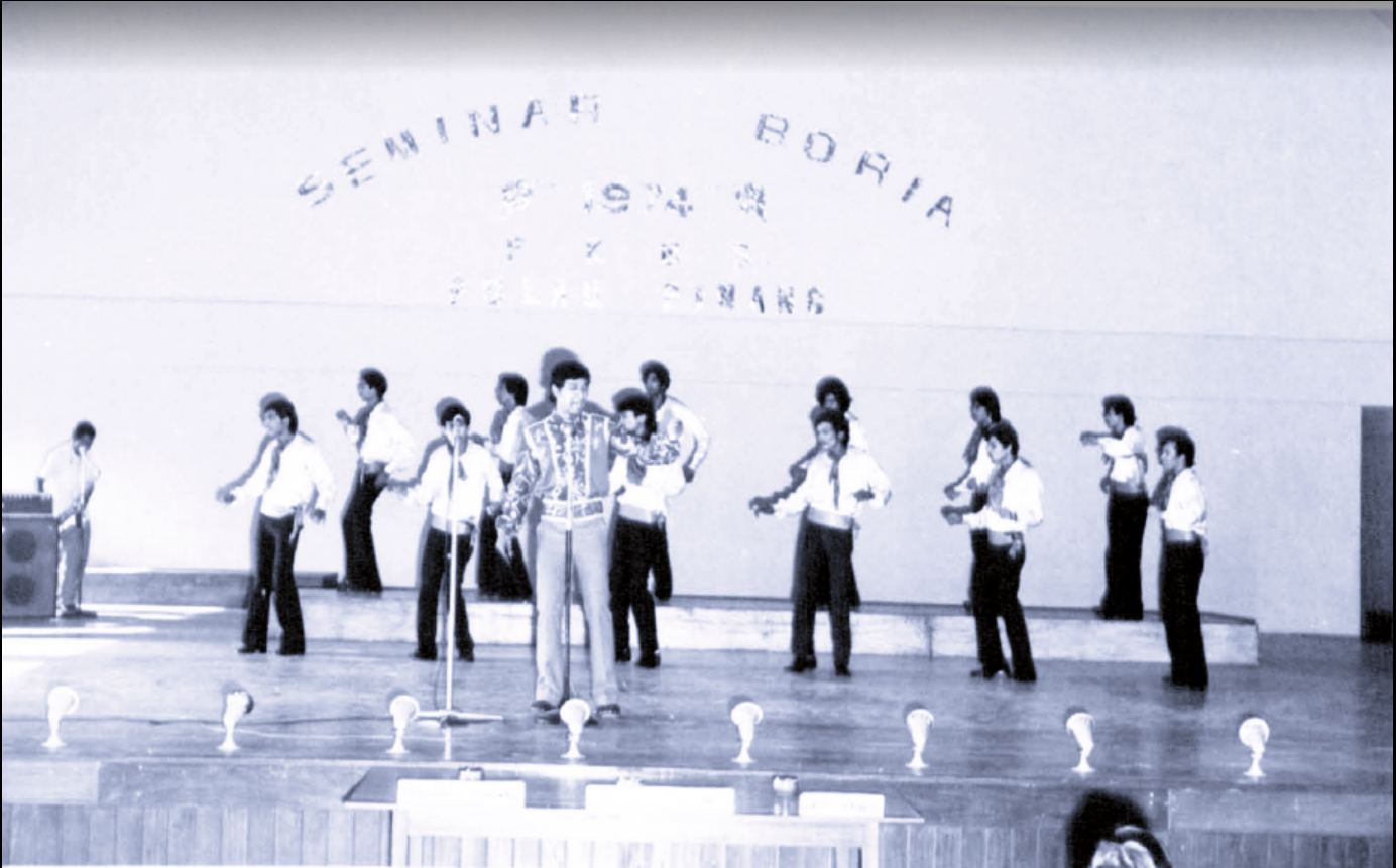 Black and white image of Boria performers. A central figure sings with a microphone while two lines of singers dance behind him.