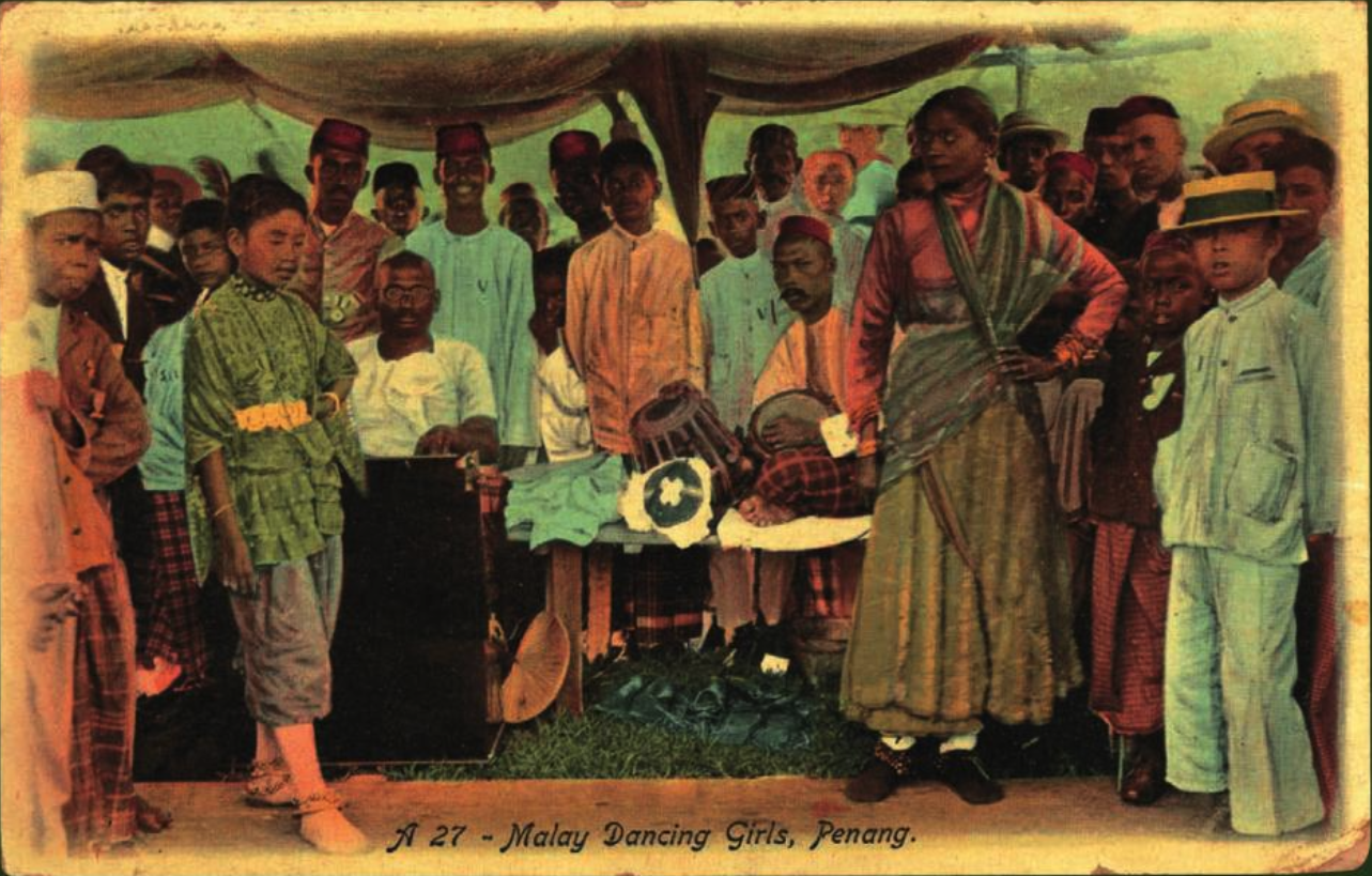 A colourised photo of two Malay 'dancing girls' surrounded by a group of boys and men.