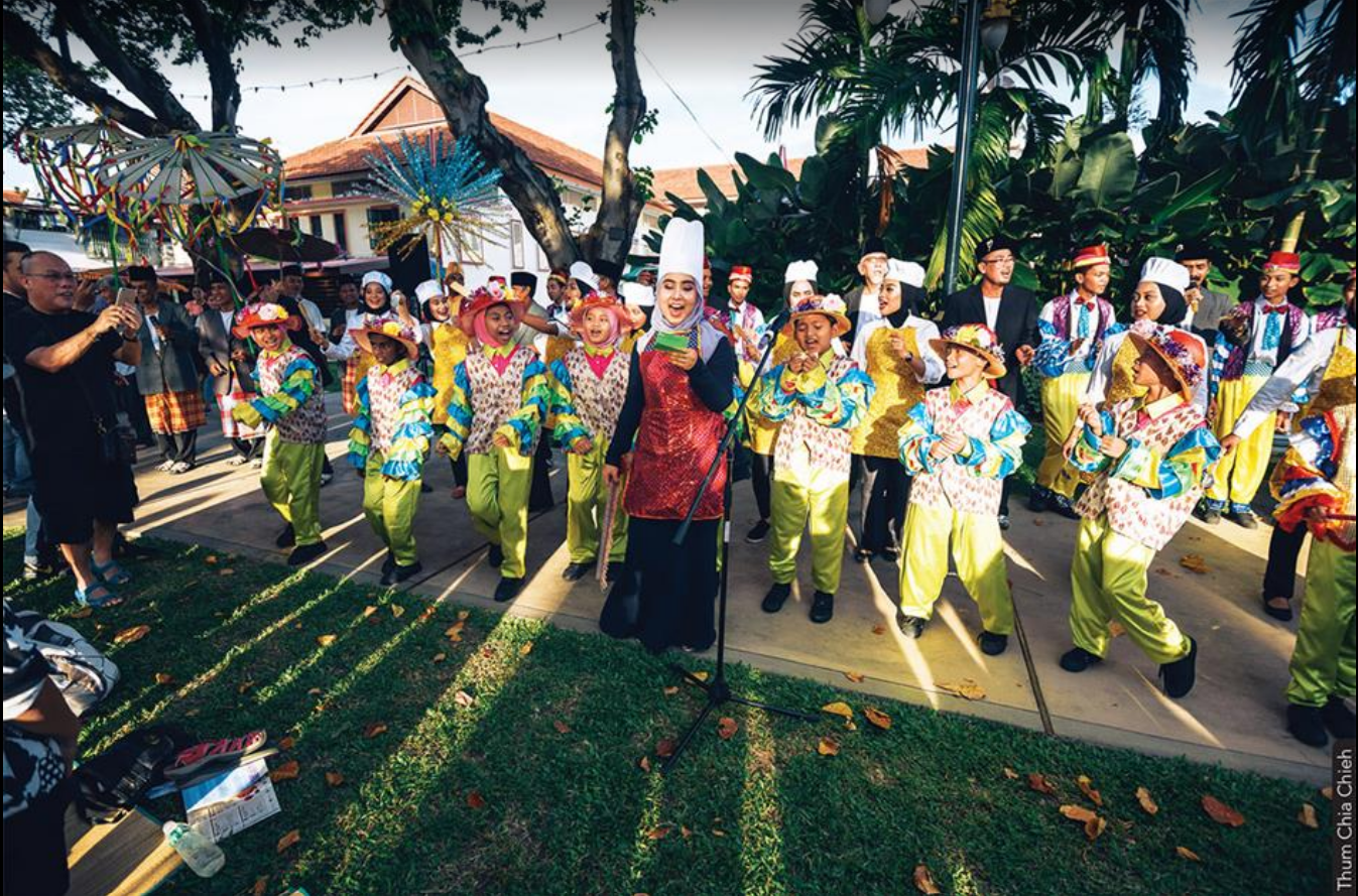 Group of children dancing and singing with female lead in a courtyard with greenery.