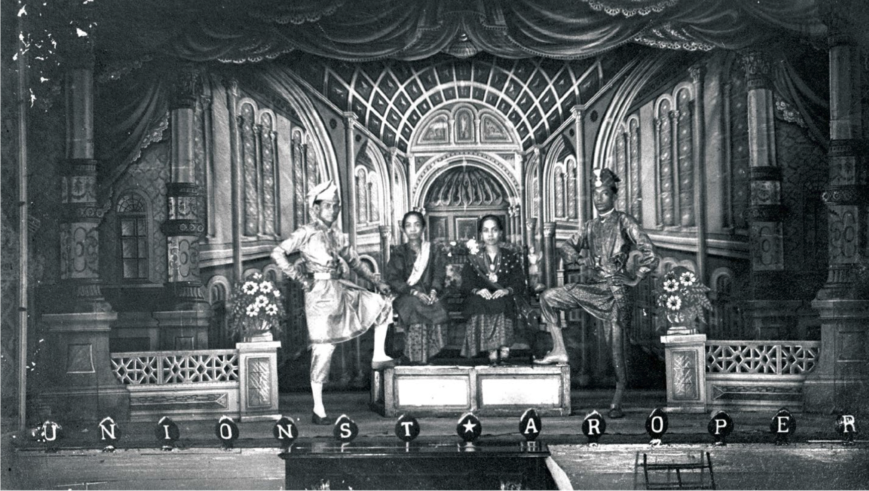 A staged photo of four people on a stage. Two women are seated and flanked by two standing men. They are in an opulent scene.