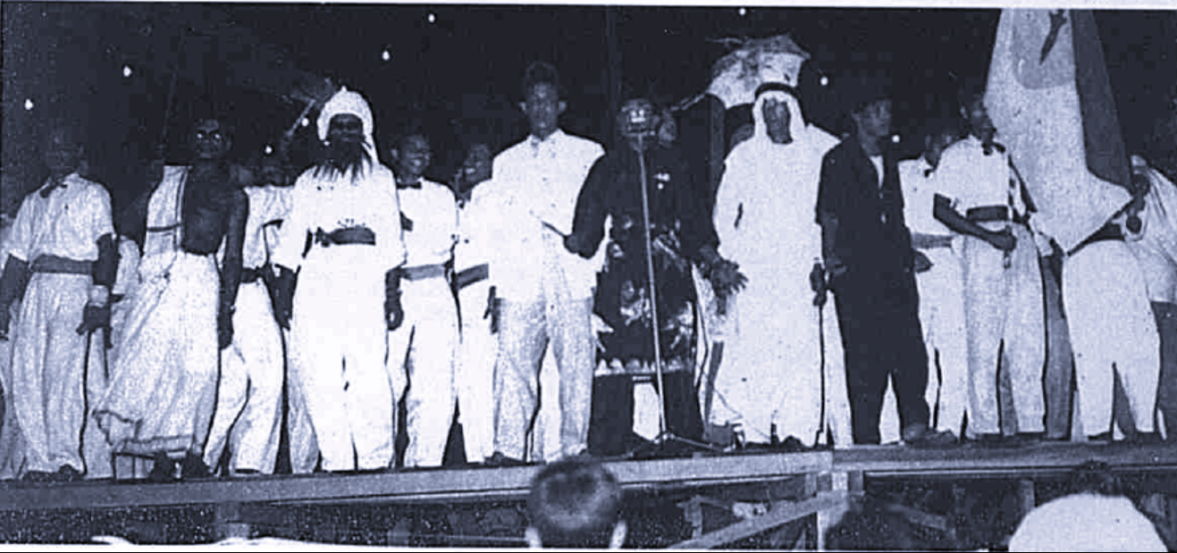 Black and white photo of performers. Man singing with microphone with rest of group behind him performing dance.