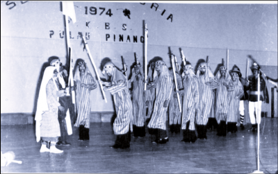 Black and white image of group in costume performing Boria, holding sticks and flags.