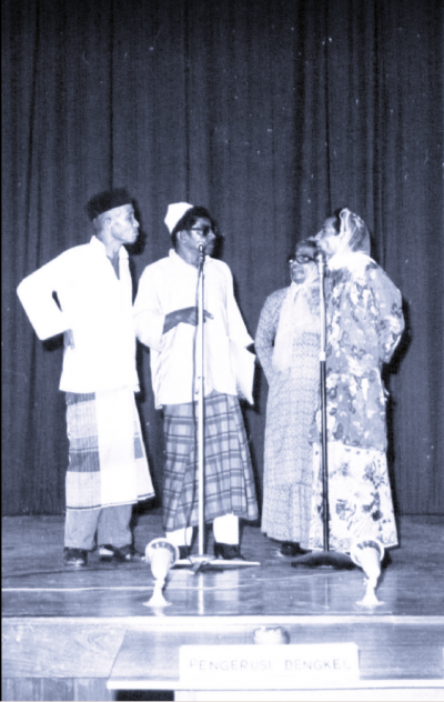 Black and white image of four people on stage with microphone. Two elderly men and two women.