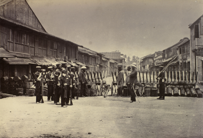 Black and white photo of Malay soldiers in formation behind a barricade. They hold rifles.