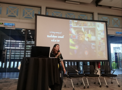 A presenter paying tribute to Roslisham Ismail, with his photo displayed on the screen behind her.